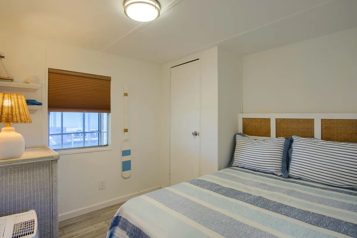 A tranquil bedroom with a queen bed featuring striped bedding, a warm-glow lamp on the nightstand, a decorative oar on the wall, and a window with blinds that lets in natural light. The soft colors and coastal accents create a peaceful vibe.