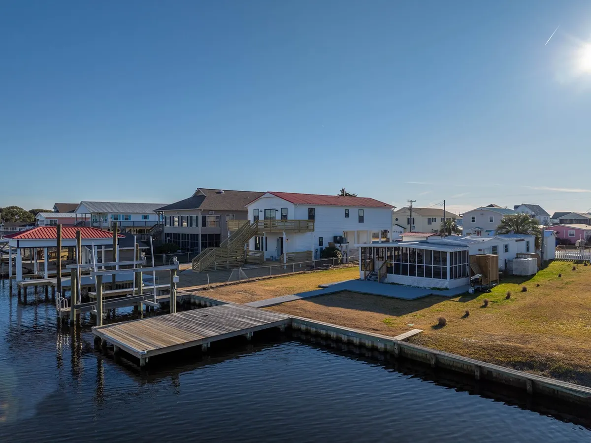 The image showcases a charming waterfront house with vibrant red roofs, set beside a serene canal. A private dock stretches out over calm water, inviting guests to relax, fish, or launch a boat. The spacious open yard offers ample room for outdoor gatherings, framed by gentle sunlight reflecting off the water and lush coastal surroundings.