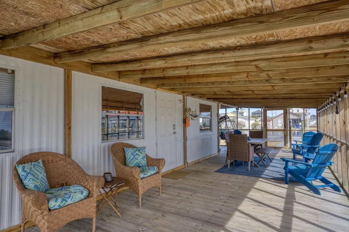 An outdoor dining area featuring wicker chairs, a table, and blue Adirondack chairs beneath wooden beams. The setting is casual and inviting, ideal for gathering with friends or family for meals or socializing. The flat deck surface is clearly visible, enhancing the sense of easy transitions from inside to outside.