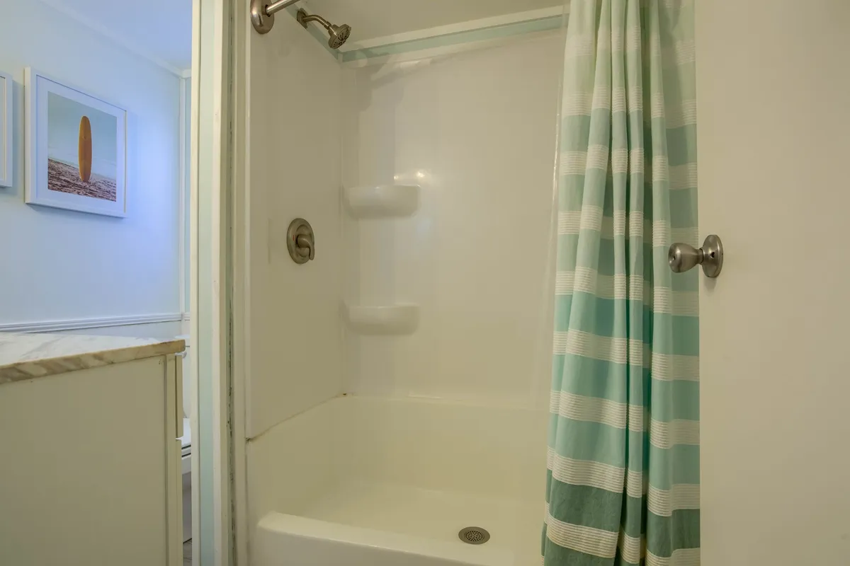 A calm, serene bathroom with a step-in shower featuring a striped curtain, light blue walls, and decorative beach-themed artwork. The space feels fresh and accessible, echoing the coastal design.