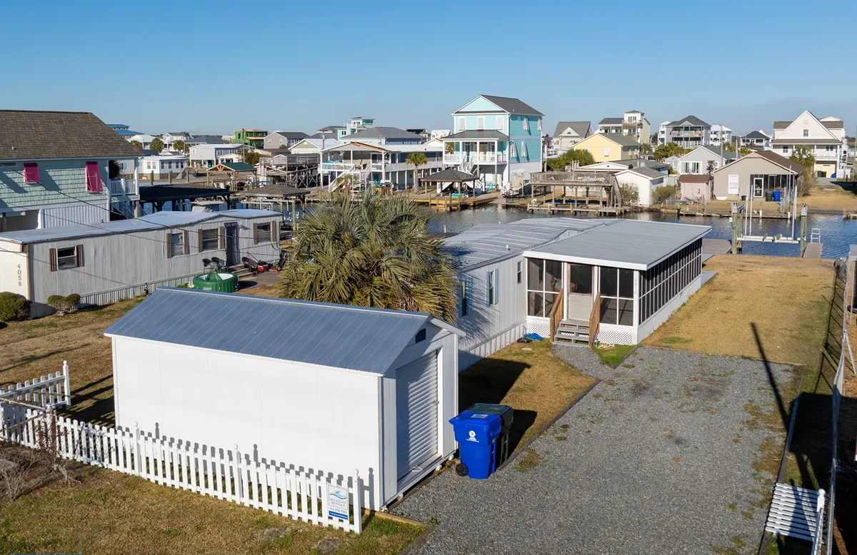 An inviting coastal exterior featuring a white shed, lush green palm tree, a screened porch perfect for relaxing, and hints of the property’s outdoor amenities. The blue trash can and casual layout evoke the laid-back atmosphere of island life, while the open outdoor space hints at easy access to local activities like boating or fishing.