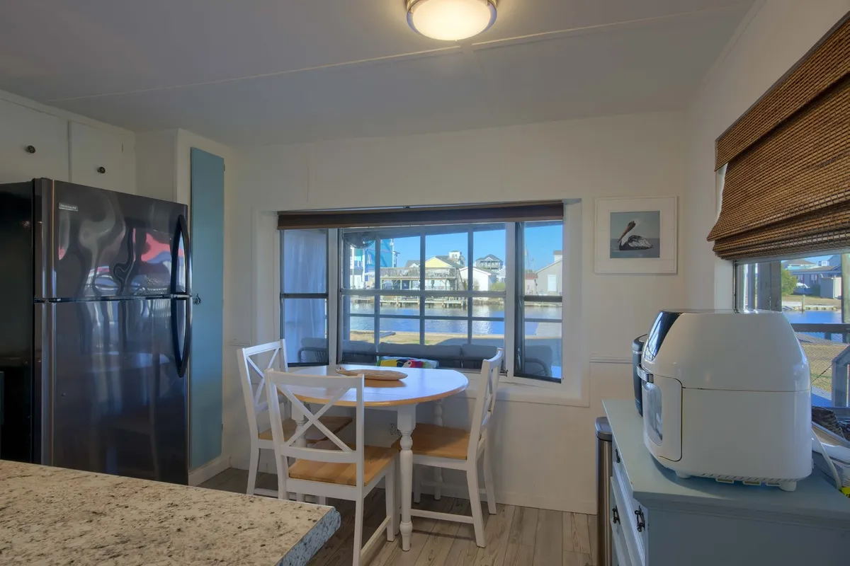 The image features a round dining table surrounded by white chairs, situated beside a large window that offers a scenic view. The bright, sunlit space is ideal for enjoying breakfast or quick meals while taking in the water views, capturing both practicality and the coastal charm mentioned in the section.