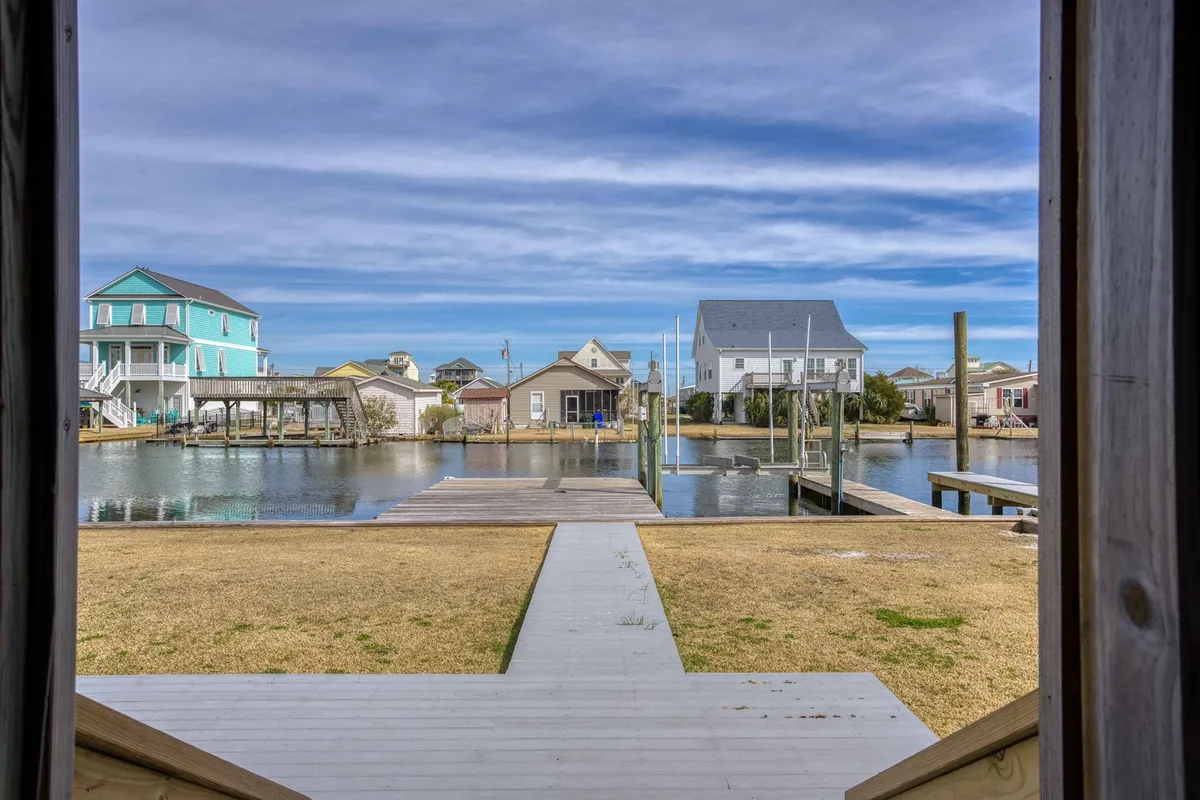 A tranquil canal-side setting with a private dock in the foreground, surrounded by lush green grass and a view of neighboring homes. The scene captures the calm water, moored boats, and ample dock space, evoking the accessibility and appeal of waterfront living. The lighting hints at either a peaceful morning or serene evening, ideal for fishing or simply relaxing by the water.