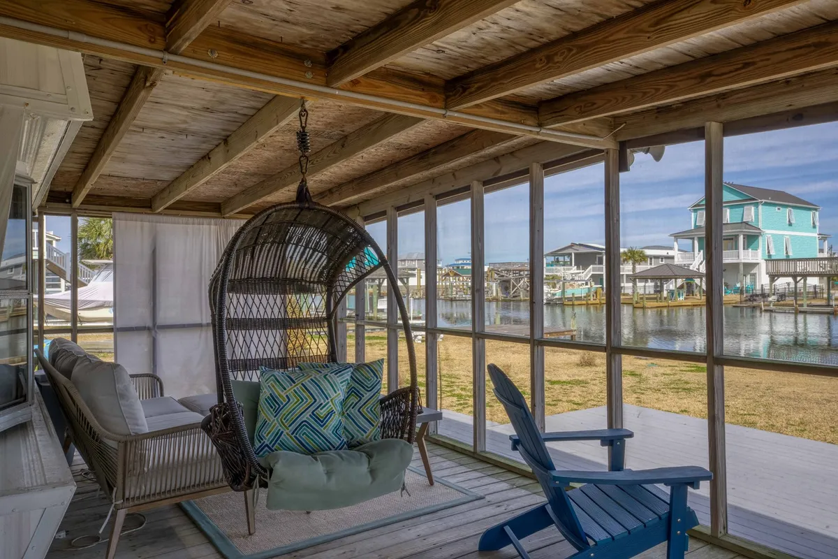 This image shows a tranquil screened porch with a hanging basket chair, sofa adorned with colorful pillows, and a beautiful view of the water beyond the porch's screens. The space is airy and inviting, perfectly blending indoor comfort with the outdoor setting.
